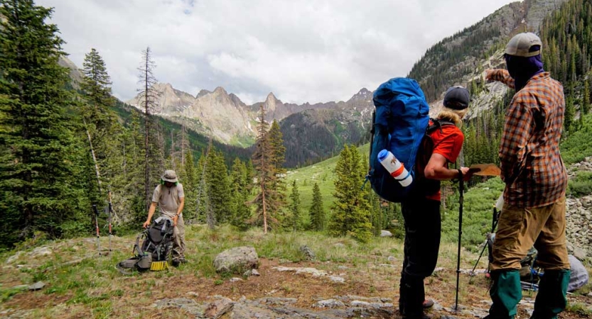 Two people look out over the mountainous landscape, while another rests with their backpack on the ground.